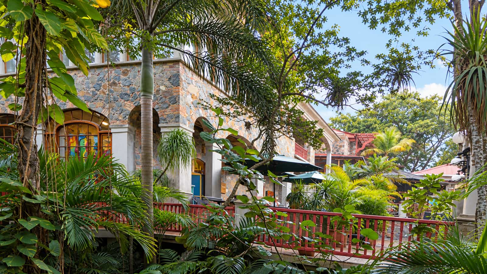 Edificios de piedra con arcos, barandillas rojas y plantas tropicales en Mongoose Junction.